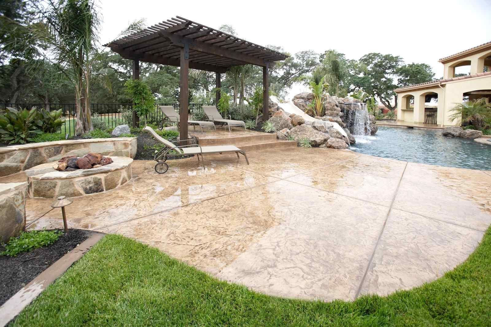 A luxurious backyard in Phoenix features a large pool with a stone waterfall, a fire pit with seating, and a shaded pergola over lounge chairs. The concrete pool deck installation by a dependable concrete installer enhances the area, which is surrounded by lush greenery and landscaped plants, creating a serene and inviting atmosphere.